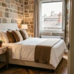 Chambre de boutique-hôtel avec lit en lin blanc, murs en pierre et vue sur les toits d'une ville européenne