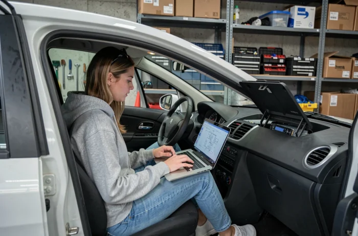 Jeune femme consultant un schéma de câblage autoradio sur ordinateur portable dans une Peugeot 207 garée dans un garage résidentiel